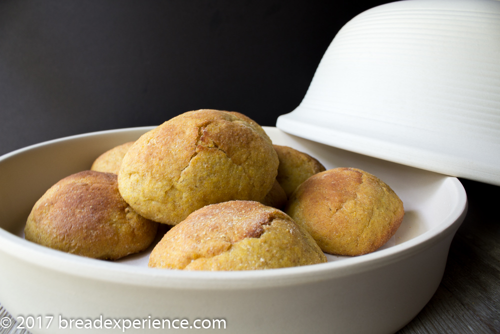 Kefir Milk Einkorn Pumpkin Cornmeal Rolls Baked in La Cloche Golden-orange pumpkin cornmeal einkorn rye rolls in a white baker with the lid resting beside them.