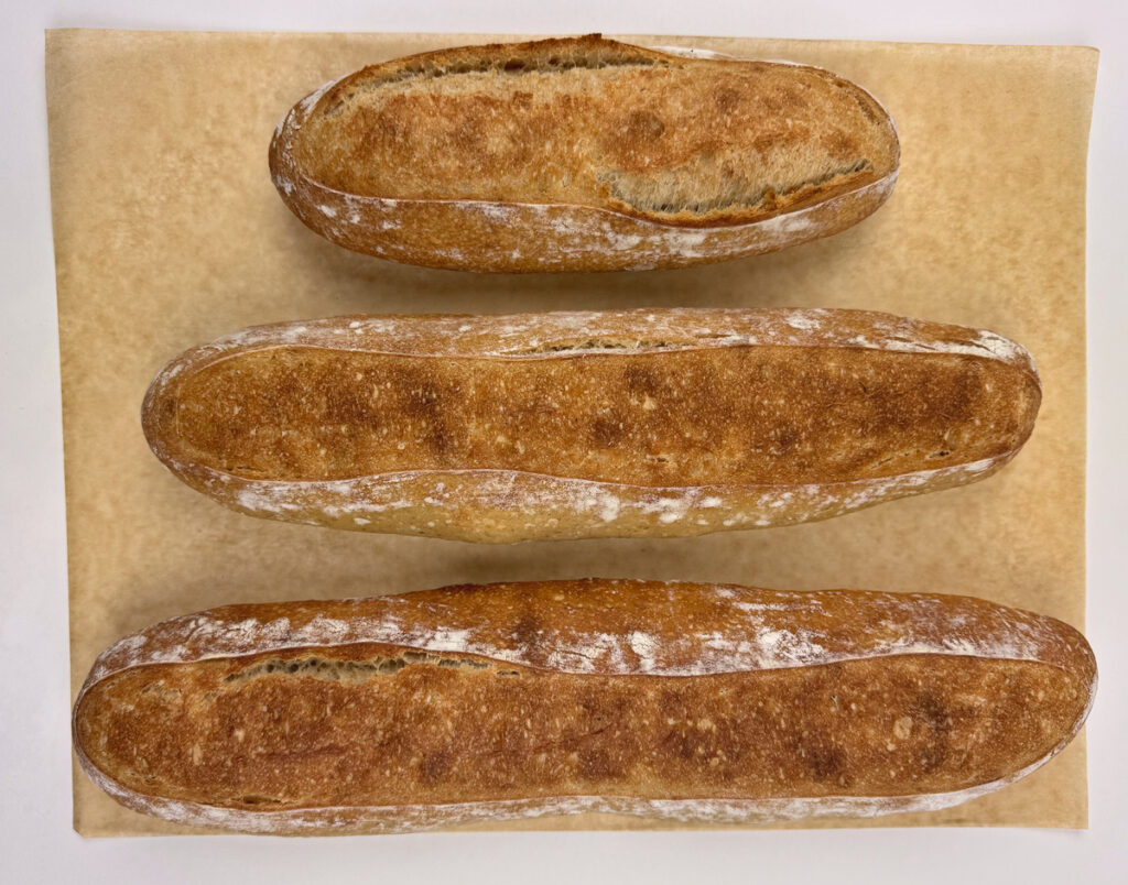 Three baked baguettes in graduated sizes on parchment: a short plump loaf, a medium, and a long generous loaf.
