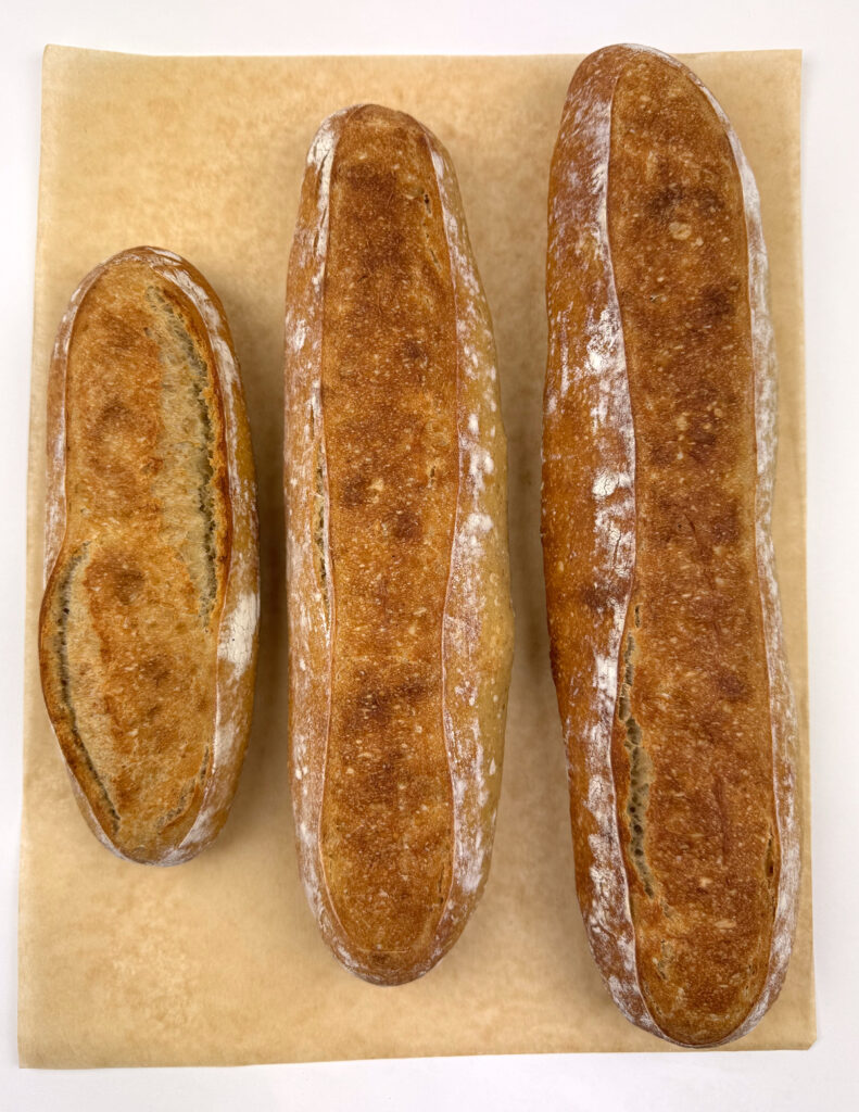 Three baked baguettes on parchment, each scored with a single center slash and baked to a deep golden, floury crust.