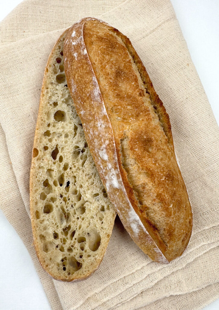 A baguette cut lengthwise showing an open, irregular crumb with large holes, beside its crisp golden crust.
