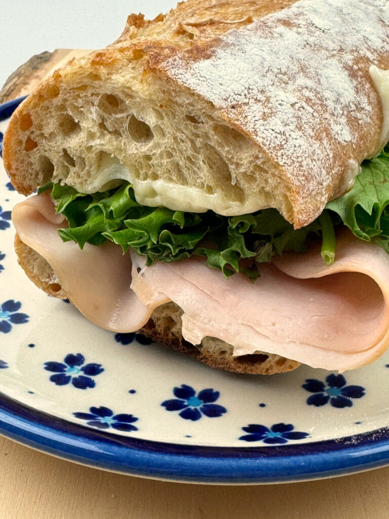 A baguette sandwich with ham, melted cheese, and green lettuce on a blue floral plate, crust dusted with flour.
