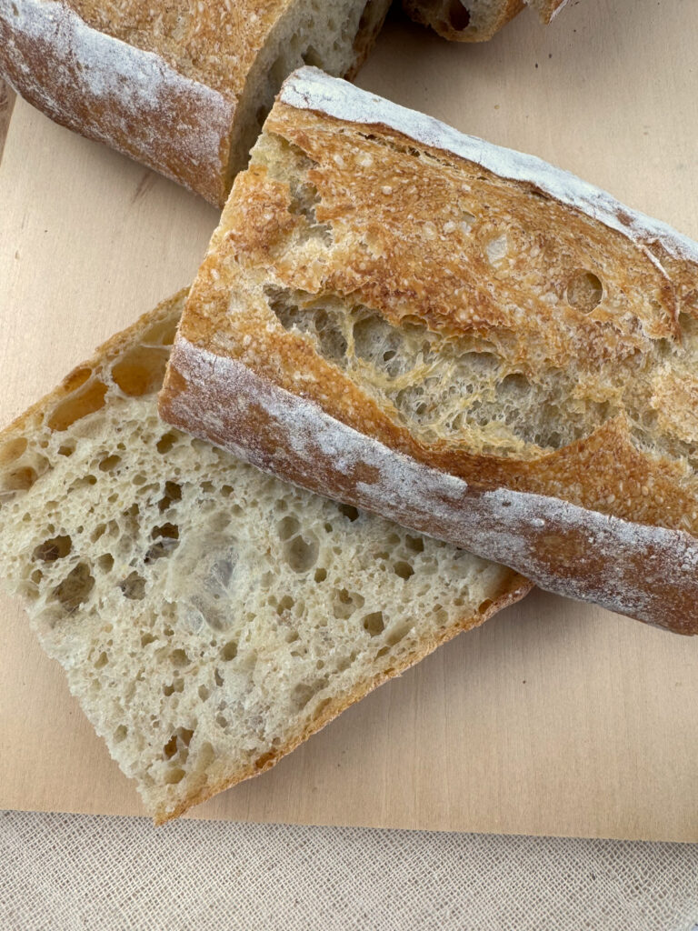 Thick slices of crusty baguette on a wood board, showing an open airy crumb and golden flour-dusted crust.