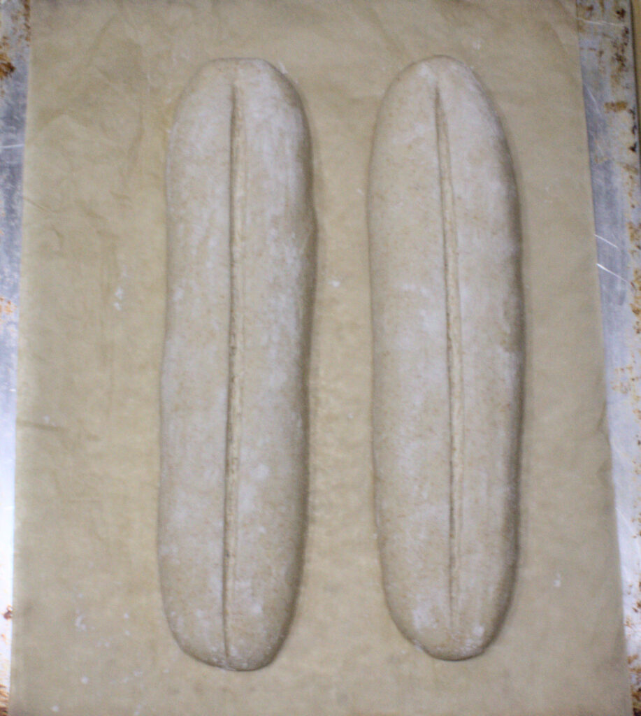 Two shaped sourdough NOLA French bread loaves on parchment paper, each scored with one long slash down the center before baking.