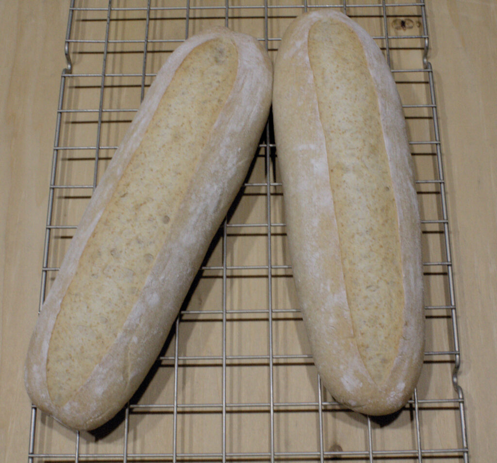 Two baked sourdough NOLA French bread loaves on a cooling rack viewed from above, with one showing an opened ear from scoring.