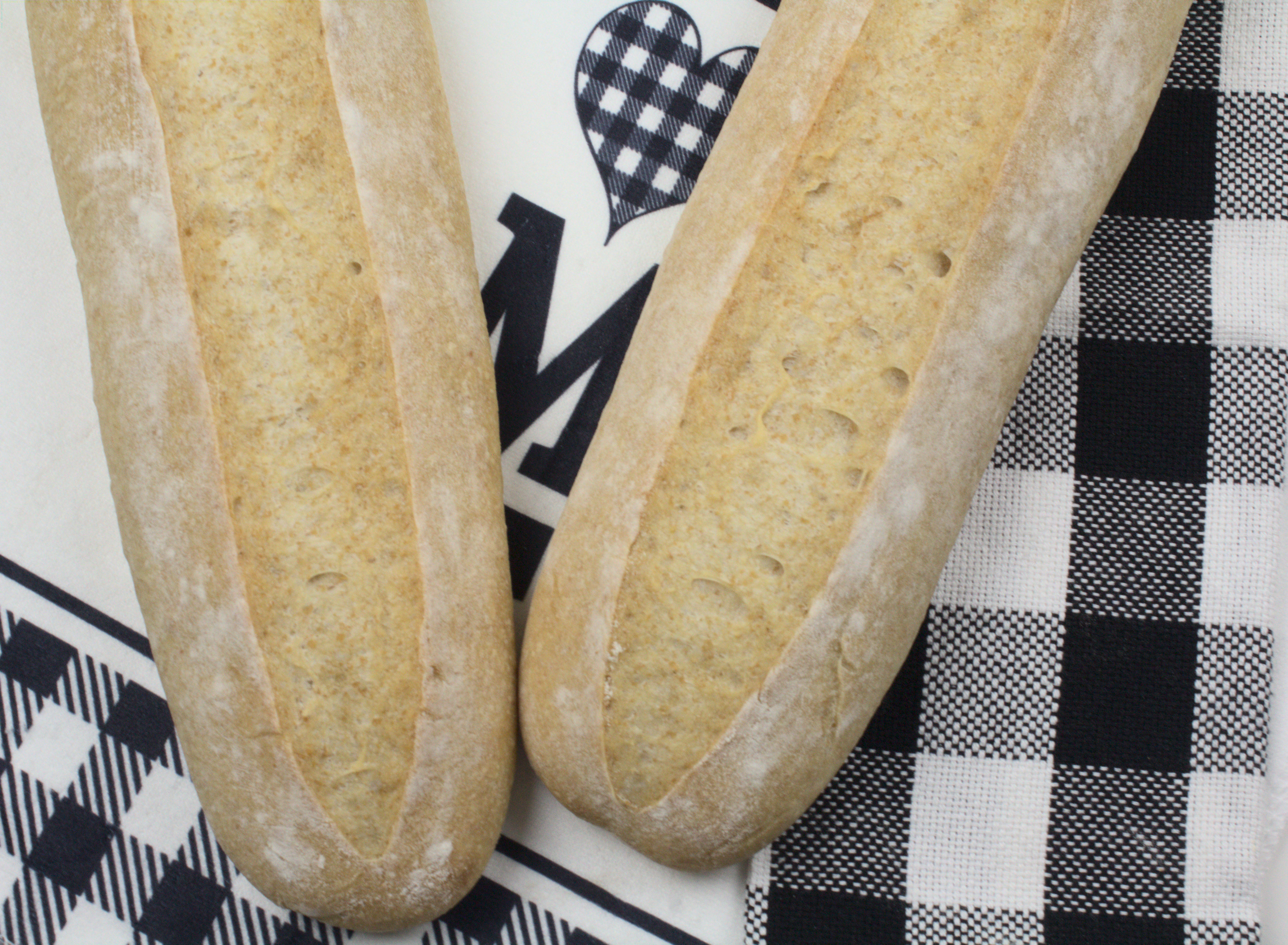 Two long oval loaves of sourdough New Orleans French bread with 25% whole grain spelt flour, viewed from above on a black and white checkered cloth, showing pale golden crusts dusted with flour.
