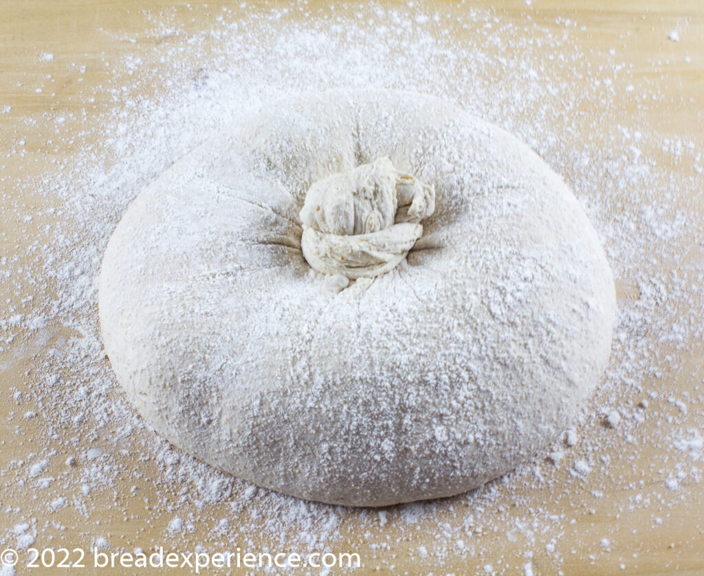 Knot on top of final bake Shaped Pan Gallego with central twisted knot, dusted with flour on a wooden surface.