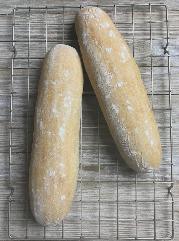 Two baked sourdough NOLA French bread loaves resting on a wire rack, demonstrating the contrast between a scored loaf with visible ear and an unscored loaf with a smoother surface.