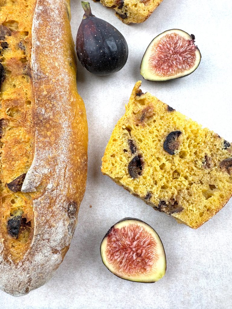 Close-up of a pumpkin fig baguette slice with a golden open crumb and dark fig pieces, beside Brown Turkey fig halves