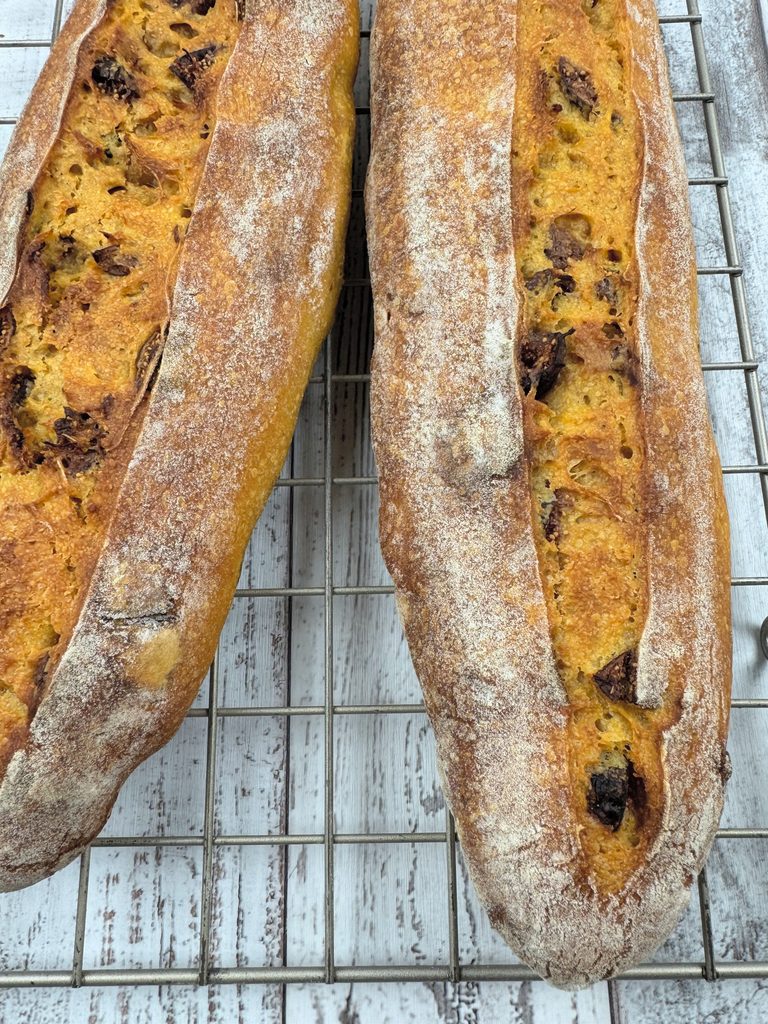 Two pumpkin fig baguettes on a cooling rack, each with one long open score showing golden crumb and figs