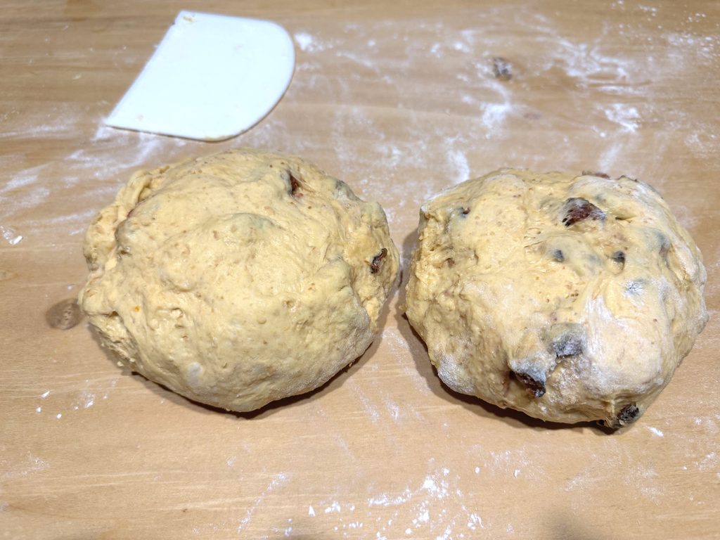 Two rounded pieces of pumpkin and fig dough on a floured wooden board beside a white bench scraper
