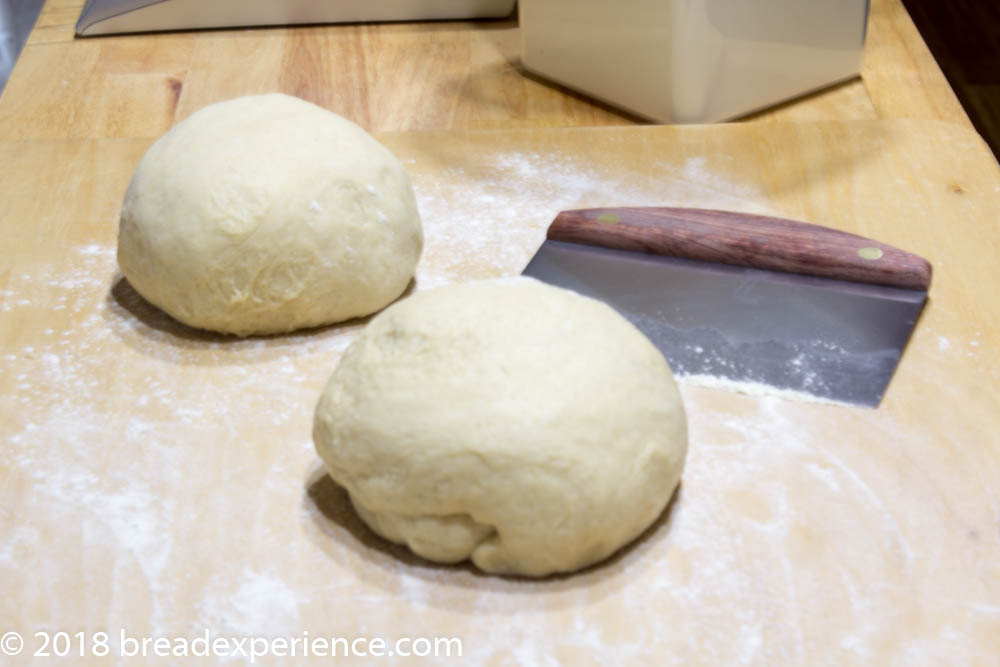 Two balls of sourdough divided on a floured wooden surface next to a metal bench scraper.