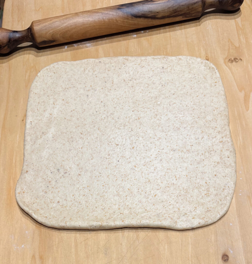 Dough rolled into a square on a wooden board, flecked with whole spelt, a rolling pin resting behind it.