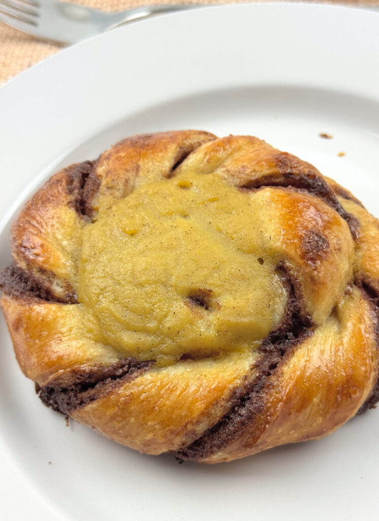 A single Sourdough Solskinnsboller on a white plate, its twisted coil framing a vanilla custard center.