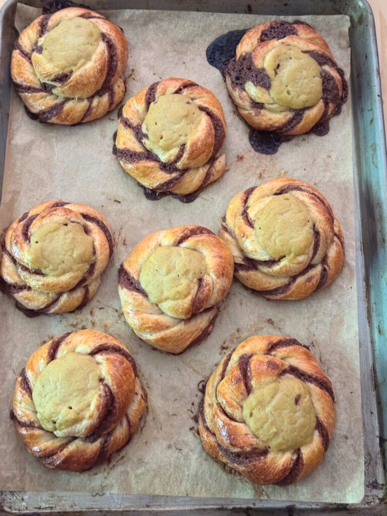 Eight Norwegian sun buns fresh from the oven on a parchment lined sheet, with cinnamon filling that escaped and baked onto the parchment beside one bun.