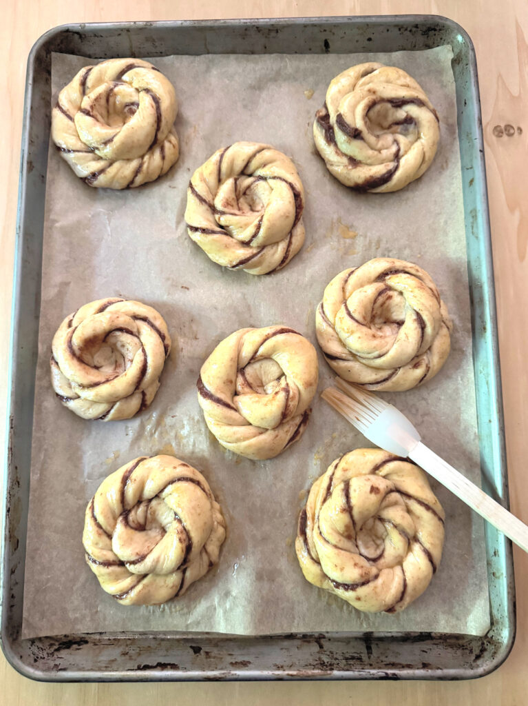 Eight sun buns brushed with egg wash for a glossy finish, a pastry brush resting on the sheet.