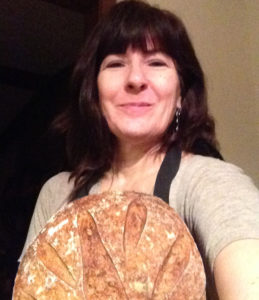 Woman with long dark hair, smiling and wearing an apron, holding a round loaf scored with a petal pattern.