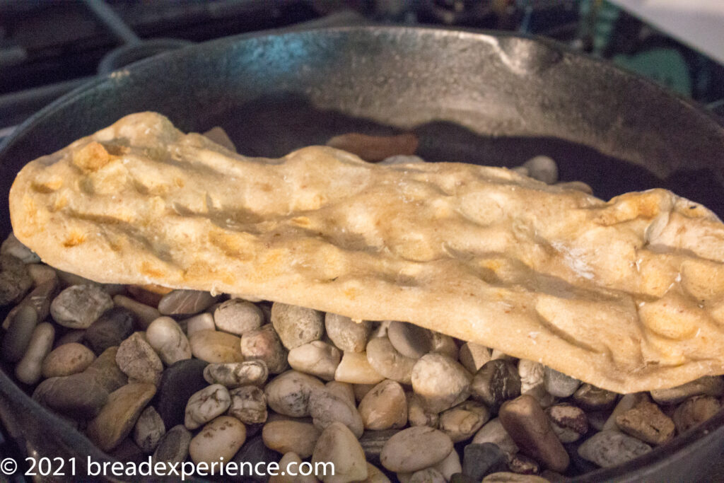 Underside of a baked Sangak flatbread resting on river rocks in a cast-iron skillet, showing the dimpled crust.