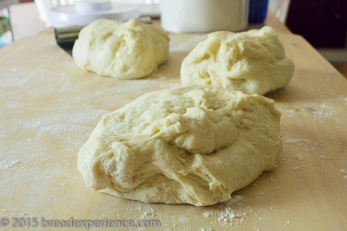 Three roughly shaped pieces of sourdough Easter braid dough resting on a lightly floured wooden work surface.